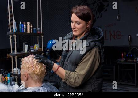 Süßes und angenehmes Mädchen, professionelle kaukasische Friseurin, trocknet die Haare einer Frau, einer erwachsenen Kundin bei der Arbeit. Stockfoto