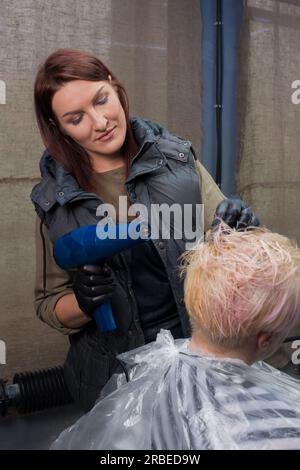 Süße und angenehme Brünette, Erwachsene Frau, europäische Friseurin, trocknet während der Arbeit die Haare eines Kunden in einem Friseursalon. Stockfoto