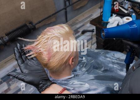 Die Barberhand eines Mädchens aus einem erfahrenen Salon, einer Mitarbeiterin in einem Handschuh, trocknet ihr Haar mit einem Fön für einen erwachsenen Kunden bei der Arbeit. Stockfoto