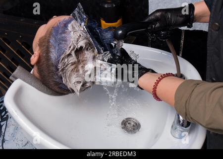 Die Hände eines professionellen Friseurs, ein Mädchen in Handschuhen, waschen sich die Farbe ab, während sie die Haare eines Klienten über einem Waschbecken mit Wasser färben, rai Stockfoto