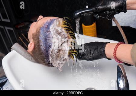 Die Hände eines professionellen Friseurmädchens bei der Arbeit mit Wasser und Regen über dem Waschbecken waschen die Farbe aus den Haaren des Klienten eines jungen Mannes während des Verfahrens ab Stockfoto