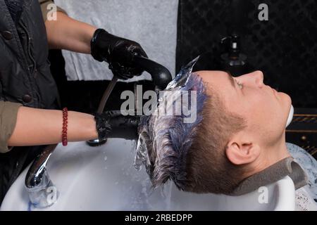 Die Hände eines erfahrenen Handwerkers, ein Mitarbeiter des Mädchensalons, waschen die Farbe ab, während sie die Haare eines jungen Mannes schneiden und färben, Stockfoto