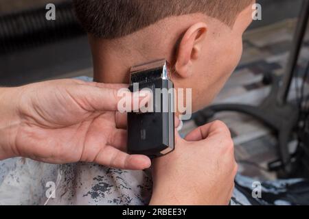 Die Hände des Friseurs haben den Hinterkopf abgeschnitten und einen modischen Haarschnitt, in der Nähe des Gehörs eines jungen Klienten mit einem speziellen Rasierer. Stockfoto