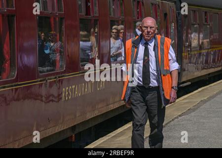 Männlicher Wachmann, ehrenamtlich und im Dienst, Gehen und vorbei an glänzenden stationären Lokomotiven (reflektierende Personen) - Haworth Station, West Yorkshire, England, Großbritannien. Stockfoto