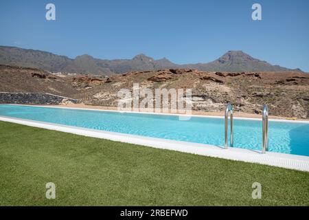 Luxuriöser langer Swimmingpool mit Blick auf die trockenen vulkanischen Berge, umgeben von einem perfekt getrimmten Rasen, ruhige Atmosphäre eines gehobenen Hauses Stockfoto