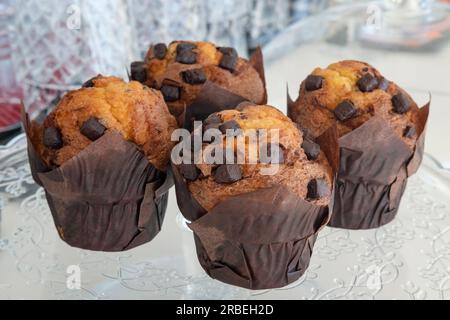 Vier köstliche, übergroße Muffins mit Schokoladensplittern, umwickelt in Pergamentpapier, ruhen auf einer großen Glasplatte, frisch zubereitetes, köstliches Dessert Stockfoto