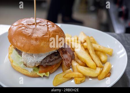 Großer Burger mit Ei und Salat, und eine große Portion frittierter Chips auf einem Teller, fertig serviert zu werden, das wegen seines Geschmacks und seiner Bequemlichkeit bevorzugt wird Stockfoto