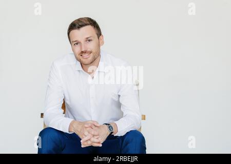 Fröhlicher Unternehmer, formelle Kleidung, sitzt auf dem Stuhl, weißer Hintergrund. Ein glücklicher Mann lächelt, freut sich über den Erfolg. Büroangestellter. Stockfoto