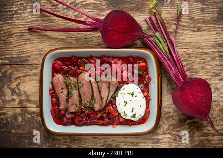 Hausgemachte Borschtsch-Suppe serviert mit gebratenen Wildscheiben aus einem Reh-Sattelfilet, serviert mit Creme Fraiche. Ausgestellt auf einer Holzboa Stockfoto