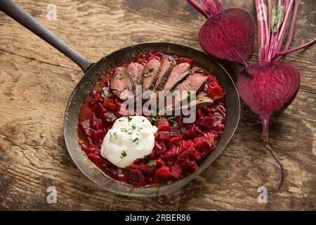 Hausgemachte Borschtsch-Suppe serviert mit gebratenen Wildscheiben aus einem Reh-Sattelfilet, serviert mit Creme Fraiche. In einer Pfanne auf einem hölzernen Cho ausgestellt Stockfoto