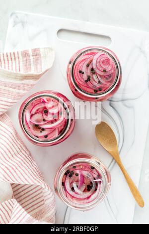 Vertikale Ansicht von drei Gläsern eingelegter roter Zwiebeln auf einem weißen Schneidebrett. Stockfoto