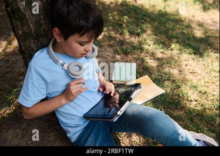 Blick von oben auf ein Schulkind, einen Jungen im Teenageralter mit Kopfhörern, der auf dem Gras im Park sitzt und Hausaufgaben auf einem digitalen Tablet macht. Kinder. Online-Lernen. Stockfoto