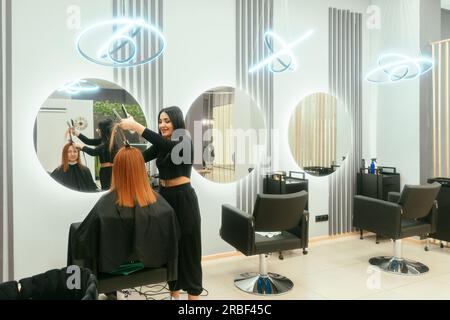Moderner, trendiger Schönheitssalon. Eine Styistin, die dem Kunden Haare und Haare frisiert. Wunderschöne orientalische Brünette arbeitet im Friseursalon. Eine Frau mit roten Haaren sitzt im Friseurstuhl und schaut in den Spiegel. Haarglättung. Stockfoto