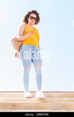 Eine junge, wunderschöne weiße Frau mit lockigem Haar, die Jeans, gelbe Bluse, Rucksack und Sonnenbrille trägt, geht eine Treppe runter, während sie ihre Intelligenz benutzt Stockfoto