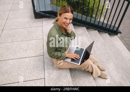 Draufsicht einer selbstständigen Frau, die auf Treppen mit einem Laptop sitzt, während sie an einem Projekt aus der Ferne arbeitet Stockfoto