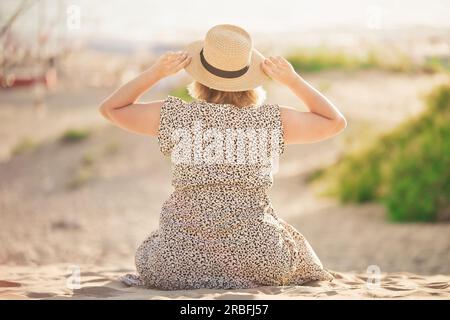 Junge Frau sitzt am Strand, hält einen Hut mit den Händen und schaut in die Ferne. Junge Frau genießt sonnigen Sommertag im Urlaub. Stockfoto