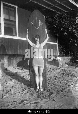 Hawaii: ca. 1917 Eine Frau und ihr langes Surfbrett an einem Strand in Hawaii. Stockfoto