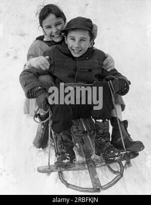 Vereinigte Staaten: c. 1955. Zwei lächelnde Kinder, die auf einem Schlitten im Schnee sitzen. Stockfoto