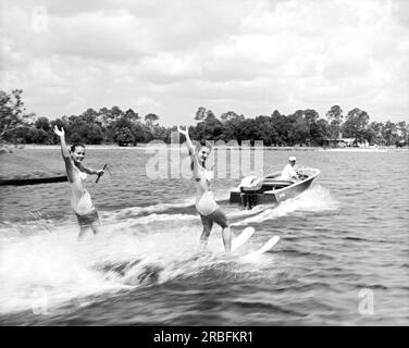 USA: ca. 1959 zwei junge Frauen, die auf einem See Wasserski fahren, winken der Kamera zu, während sie vorbeifahren. Stockfoto