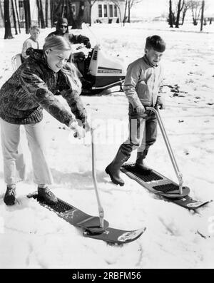 Vereinigte Staaten: c. 1967 Ein Junge und ein Mädchen scheinen sich nicht sicher zu sein, ob sie das neue Lenkskierbrett namens Fun-Ski ausprobieren wollen. Stockfoto