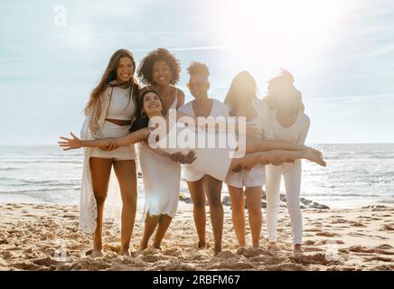 Aufgeregte weibliche Freunde feiern eine Hühnerparty und tragen ihre Freundin Braut am Strand Stockfoto