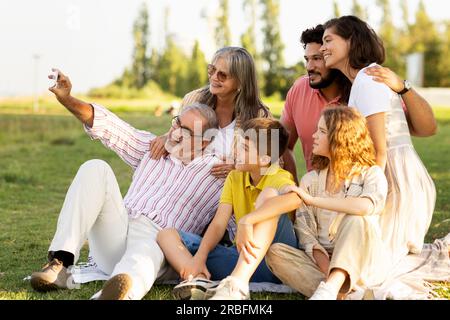 Lächelnde europäische Alte, Millennials und Teenager haben Spaß beim Picknick und machen Selfie auf dem Smartphone im Park Stockfoto