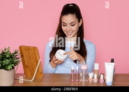 Wunderschöne junge hispanoamerikanerin, die am Tisch sitzt und Gesichtscreme benutzt Stockfoto
