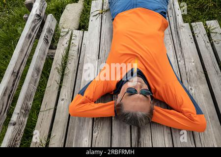 Ein Mann mit Sonnenbrille liegt auf einem Holztisch und sieht in den Himmel Stockfoto