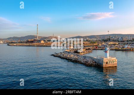 Der Eingang zum Hafen von Piräus in Griechenland, mit dem Wellenbrecher und der Stadt Piräus mit Fabrikschornsteinen im Hintergrund. Stockfoto