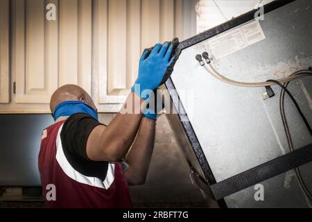 Ethnischer Auslieferer mit Maske neigt alten Kühlschrank auf Transportwagen mit Küchenschränken im Hintergrund - Nahaufnahme und selektiver Fokus Stockfoto