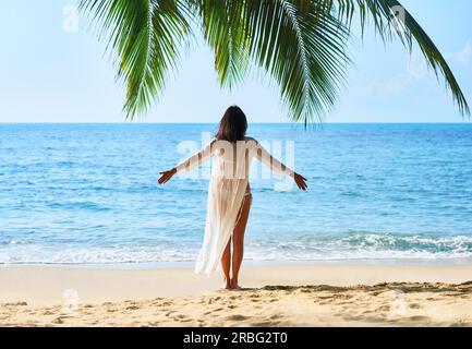 Rückansicht des freien junge Frau entspannen Sie sich und genießen Sie das Meer stehen unter Palmen am tropischen Strand. Sommer Urlaub Konzept Stockfoto