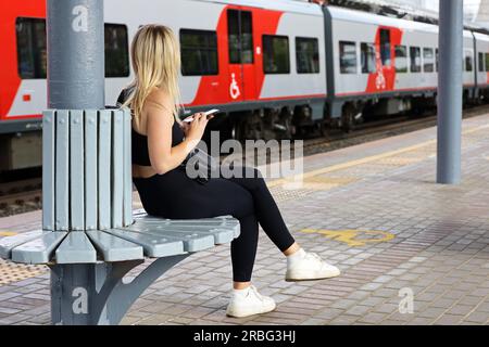 Blondes Mädchen, das auf den ankommenden Zug wartet, sitzt mit Smartphone in der Hand am Bahnhof. Pendlerzugpassagier, Reise im Sommer Stockfoto