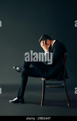 Traurig, unglücklich Mann im schwarzen Anzug auf dem Stuhl und geschlossene Fläche durch zwei Hände. Profil ansehen Portrait. Depression und Schmerz Konzept Stockfoto