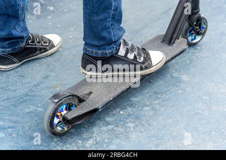 Die Beine des Fahrerjungen werden in Sneakern auf dem Stunt-Roller auf der Skate-Park-Rampe zusammengezogen. Ein Junge auf einem Roller in einem Skatepark spielt Sprünge und Tricks. Extreme Cit Stockfoto