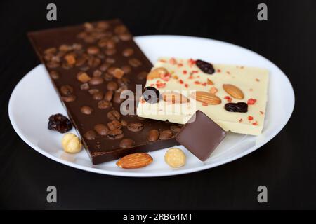 Weiße und dunkle Schokolade mit getrockneten Rosinen, Kaffeebohnen und Nüssen. Leckeres Dessert auf einem Teller aus dunklem Holz Stockfoto