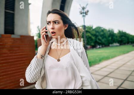 Schockierte, erstaunliche Frau, die im Hintergrund der Sommerstadtstraße auf dem Handy spricht. Wow-Konzept Stockfoto
