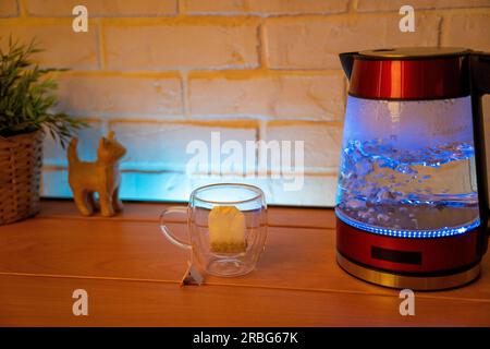 Ein elektrischer Wasserkocher aus Glas auf einem Holztisch und ein durchsichtiger Becher mit einer Tüte köstlichem Tee. Hochwertiges Foto Stockfoto