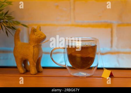 Ein elektrischer Wasserkocher aus Glas auf einem Holztisch und ein durchsichtiger Becher mit einer Tüte köstlichem Tee. Hochwertiges Foto Stockfoto