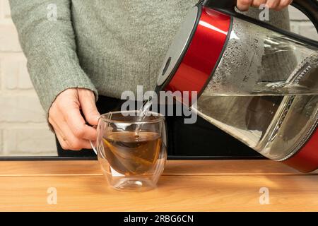Frau, die heißes, kochendes Wasser aus einem elektrischen Wasserkocher in einen durchsichtigen Glasbecher auf dem Tisch portiert. Der Prozess der Teezubereitung oder Teezeremonie in einem warmen, weichen Licht. M Stockfoto