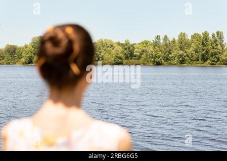 Eine Frau mit einem Chignon, die in der Nähe des Dnieper-Flusses in Kiew, Ukraine, steht, beobachtet die Bäume in der Ferne. Die Silhouette der Dame ist es Stockfoto