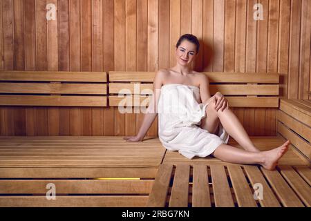 Seite Ansicht in voller Länge Portrait von junge Frau lächelnd auf Holzsofa Bank Sauna mit Haaren Donut Dutt in weißes Handtuch gewickelt Stockfoto