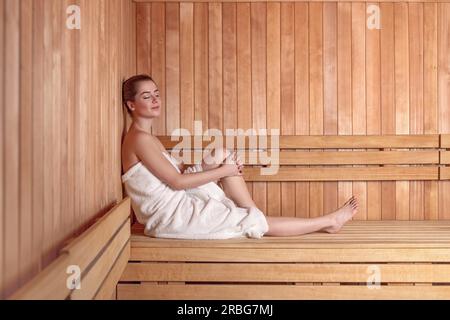 Seite Ansicht in voller Länge Portrait von junge Frau lächelnd auf Holzsofa Bank Sauna mit Haaren Donut Dutt in weißes Handtuch gewickelt Stockfoto