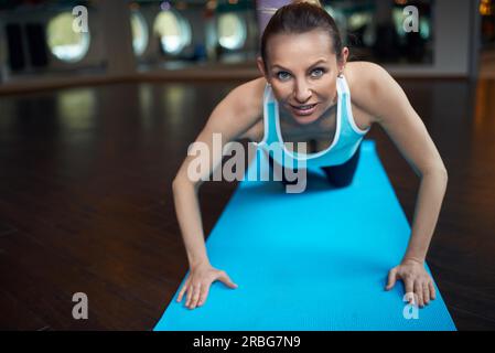 Blonde Frau mittleren Alters Liegestütze trainieren im Fitness-Studio und Blick in die Kamera beim liegen auf blaue Yoga Teppich über Spiegel Hintergrund Stockfoto