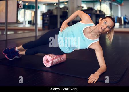 Eine Frau in einem ärmellosen Hemd und schwarzen Leggings im Fitnessstudio, die mit Schaum oder Faszie-Rollen trainiert und in die Kamera schaut Stockfoto