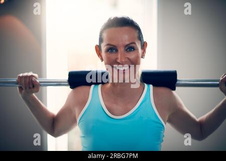 Eine attraktive blonde Frau in einem ärmellosen Hemd, die im Fitnessstudio steht, die Barbell auf den Schultern hält und in die Kamera schaut Stockfoto