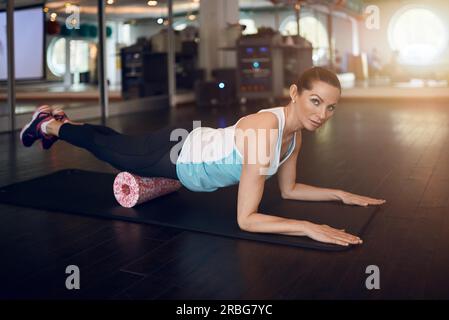 Eine Frau in einem ärmellosen Hemd und schwarzen Leggings im Fitnessstudio, die mit Schaum oder Faszie-Rollen trainiert und in die Kamera schaut Stockfoto