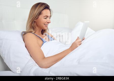 Attraktive Frau entspannen in komfortablen neutral weiß Bett mit einem Tablet-PC in ihren Händen und einem freundlichen Lächeln Stockfoto