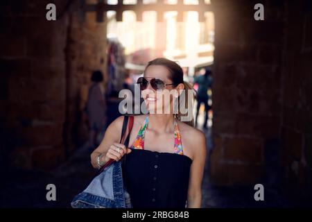 Attraktive, trendige Frau mit Sonnenbrille und einer großen Handtasche über der Schulter zum Einkaufen in der Stadt, die mit einem Lächeln zur Seite blickt Stockfoto