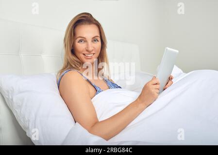 Attraktive Frau entspannen in komfortablen neutral weiß Bett mit einem Tablet-PC in ihren Händen und einem freundlichen Lächeln Stockfoto