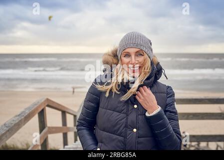Eine Frau mittleren Alters, die an einem kalten Wintertag am Meer auf einer Holzterrasse mit Blick auf den Strand stand, an einem luftigen Tag, an dem sie glücklich lächelte Stockfoto
